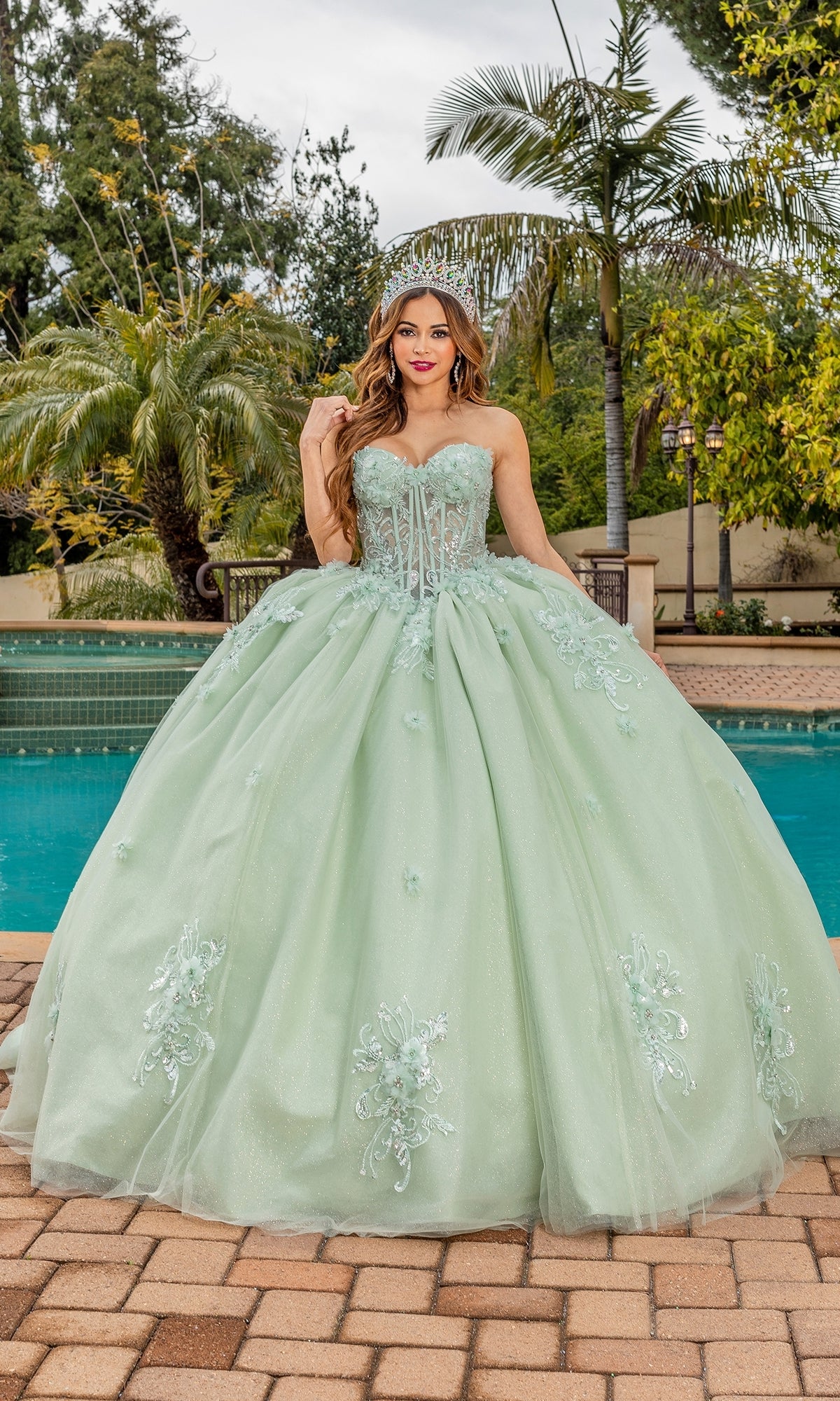 A young woman stands by a pool surrounded by palm trees, wearing the Quinceanera Dress 1893 By Dancing Queen in sage green with a strapless sweetheart neckline.